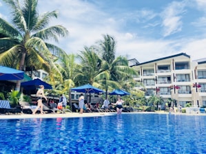 A vibrant poolside scene with people lounging and walking nearby, surrounded by tall palm trees. The setting includes a large, clear blue swimming pool with blue umbrellas lining the poolside. A modern hotel building with balconies is visible in the background under a partly cloudy sky.