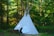 A white teepee stands in a lush, green forest. The structure is made of fabric supported by tall wooden poles and is surrounded by dense trees and foliage. Sunlight filters through the leaves, creating a serene and peaceful environment.