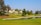 A residential area with houses lined up next to a lush green golf course. The scene includes a small water body reflecting the clear blue sky. Several tall trees, including some palm trees, are scattered around the area.