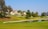 A residential area with houses lined up next to a lush green golf course. The scene includes a small water body reflecting the clear blue sky. Several tall trees, including some palm trees, are scattered around the area.