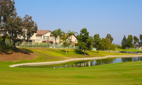 A residential area with houses lined up next to a lush green golf course. The scene includes a small water body reflecting the clear blue sky. Several tall trees, including some palm trees, are scattered around the area.