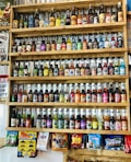 A wooden shelf filled with a large assortment of colorful glass bottles of soda. The bottles vary in label design, flavor, and color. Below the shelves, there are bags of candy and novelty items hung on display. The setting suggests a vintage or retro theme with nostalgic signage decorating the wall.