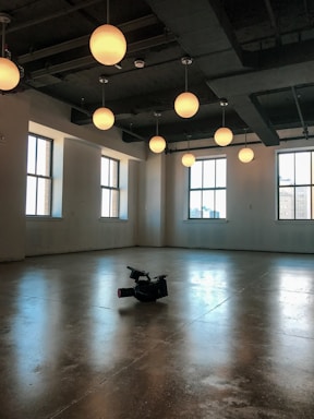 An empty room with polished concrete floors illuminated by several round ceiling lights. Large windows with natural light are visible on two walls, and a professional video camera is placed on the floor.