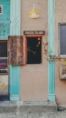 A rustic storefront with a sign reading 'Tacos To Go' above a small serving window. The building features textured beige walls and turquoise architectural accents, along with a single yellow outdoor light fixture. A wooden shutter is partially open, revealing a dark interior lit by a few warm bulbs. A note on the wall advises 'Quiet Please, Neighbors above.' A planter box with vines hangs nearby.