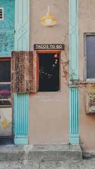 A rustic storefront with a sign reading 'Tacos To Go' above a small serving window. The building features textured beige walls and turquoise architectural accents, along with a single yellow outdoor light fixture. A wooden shutter is partially open, revealing a dark interior lit by a few warm bulbs. A note on the wall advises 'Quiet Please, Neighbors above.' A planter box with vines hangs nearby.
