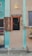 A rustic storefront with a sign reading 'Tacos To Go' above a small serving window. The building features textured beige walls and turquoise architectural accents, along with a single yellow outdoor light fixture. A wooden shutter is partially open, revealing a dark interior lit by a few warm bulbs. A note on the wall advises 'Quiet Please, Neighbors above.' A planter box with vines hangs nearby.