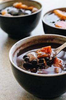 A bowl filled with a dark liquid, containing visible black tapioca pearls and chunks of sweet potato. Another bowl with similar contents is visible in the background.