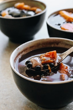 A bowl filled with a dark liquid, containing visible black tapioca pearls and chunks of sweet potato. Another bowl with similar contents is visible in the background.