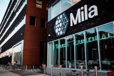 A modern building with a brick facade featuring the word 'Mila' in large letters. The windows display neon signs, and several bicycles are parked in front of the building. A person is adjusting one of the bicycles.