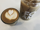 Two beverages are placed on a white surface: a hot coffee in a glass cup with intricate latte art resembling a heart, and a larger glass filled with iced coffee, bearing the letters 'FOAM' on its side.