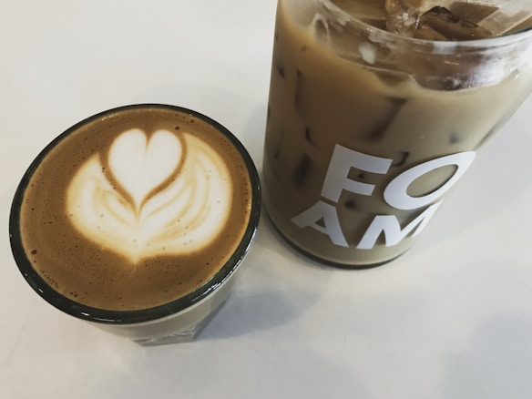 Two beverages are placed on a white surface: a hot coffee in a glass cup with intricate latte art resembling a heart, and a larger glass filled with iced coffee, bearing the letters 'FOAM' on its side.