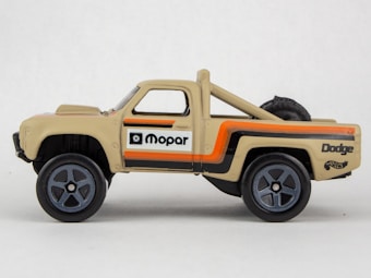 A beige toy truck with black wheels is seen. It has detailed decals on its sides including the text 'Mopar' and 'Dodge' with orange and black stripes. The model appears to be a miniature version of a pickup truck, and there is a faux spare tire mounted in the bed.