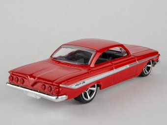 A red toy car model with a sleek design, detailed with chrome accents and black-tinted windows. The car is shown from a rear-side angle, highlighting its smooth curves and retro styling. It is set against a plain white background, emphasizing the vibrant color of the model.