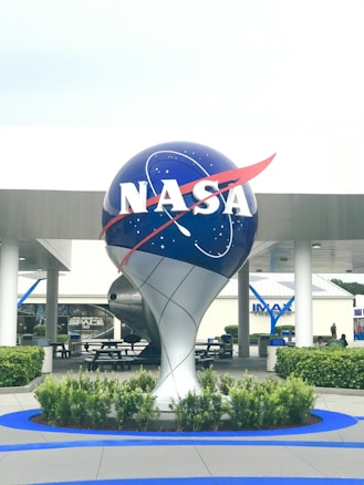 A large, decorative NASA globe with the agency's iconic logo and a starry backdrop is prominently displayed outside a modern building. Surrounding the globe are neatly arranged greenery and well-maintained bushes. In the background, a building with 'IMAX THEATER' signage is visible. The area is designed to be inviting with outdoor tables and benches.
