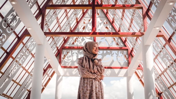 A woman wearing a floral dress and a hijab stands in an architectural space characterized by intricate lattice patterns and red beams. The ceiling is transparent, allowing natural light to illuminate the area. The expression on her face reflects contentment, and the shadows create decorative patterns on the surfaces.