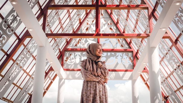 A woman wearing a floral dress and a hijab stands in an architectural space characterized by intricate lattice patterns and red beams. The ceiling is transparent, allowing natural light to illuminate the area. The expression on her face reflects contentment, and the shadows create decorative patterns on the surfaces.