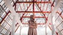 A woman wearing a floral dress and a hijab stands in an architectural space characterized by intricate lattice patterns and red beams. The ceiling is transparent, allowing natural light to illuminate the area. The expression on her face reflects contentment, and the shadows create decorative patterns on the surfaces.