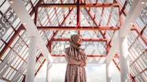 A woman wearing a floral dress and a hijab stands in an architectural space characterized by intricate lattice patterns and red beams. The ceiling is transparent, allowing natural light to illuminate the area. The expression on her face reflects contentment, and the shadows create decorative patterns on the surfaces.