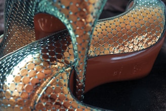 Metallic high heels with a textured, scale-like pattern featuring iridescent colors. The shoes have a gleaming surface that reflects light, showcasing a blend of teal and gold hues. The soles are a contrasting red tone, adding a distinct visual element.