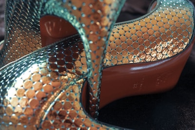Metallic high heels with a textured, scale-like pattern featuring iridescent colors. The shoes have a gleaming surface that reflects light, showcasing a blend of teal and gold hues. The soles are a contrasting red tone, adding a distinct visual element.