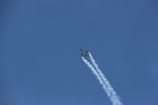 A jet aircraft is flying upwards through a clear blue sky, leaving a trail of white vapor behind it. The plane appears sleek and aerodynamic, suggesting high speed and advanced engineering.