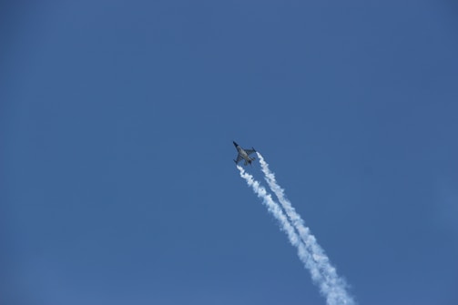 A jet aircraft is flying upwards through a clear blue sky, leaving a trail of white vapor behind it. The plane appears sleek and aerodynamic, suggesting high speed and advanced engineering.