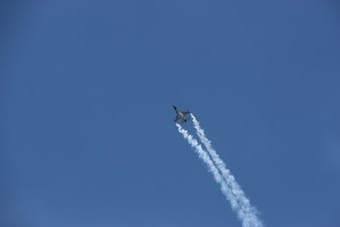 A jet aircraft is flying upwards through a clear blue sky, leaving a trail of white vapor behind it. The plane appears sleek and aerodynamic, suggesting high speed and advanced engineering.