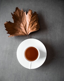 A cup of tea sits on a white saucer against a gray surface, accompanied by a large, dried brown leaf placed above it.