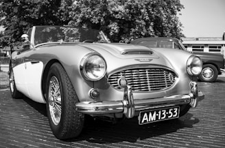 A classic convertible car is parked on a street. The vehicle features chrome accents and a distinctive front grille. The license plate reads 'AM-13-53'. Behind it, another vintage vehicle is partially visible. Trees and a building are in the background, indicating an outdoor setting.
