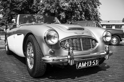 A classic convertible car is parked on a street. The vehicle features chrome accents and a distinctive front grille. The license plate reads 'AM-13-53'. Behind it, another vintage vehicle is partially visible. Trees and a building are in the background, indicating an outdoor setting.