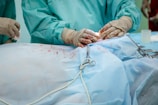 Medical professionals wearing surgical gowns and gloves are performing a surgical procedure. Various surgical instruments, including clamps, are visible on a surgical drape, which has blood stains on it.