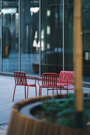 Red metal chairs are arranged in an outdoor setting with a modern glass building in the background. A large, round planter with greenery is in the foreground, adding a touch of nature to the urban environment.