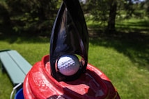 A golf ball is placed on top of a red container with a black, curved handle extending upwards. In the background, there is a grassy area and some trees, indicating an outdoor setting. The golf ball has branding on it, and the sun casts shadows on the scene.