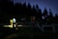 A group of people is gathered outdoors at dusk or nighttime, some holding flashlights. They stand on a grassy area near a wooden fence, surrounded by tall coniferous trees.