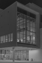 A modern building facade with large glass windows reflecting the interior lighting. The structure features geometric shapes and lines, creating a sense of order and symmetry. The interior is visible through the windows, with silhouetted figures and minimalist decor.
