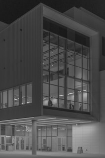 A modern building facade with large glass windows reflecting the interior lighting. The structure features geometric shapes and lines, creating a sense of order and symmetry. The interior is visible through the windows, with silhouetted figures and minimalist decor.