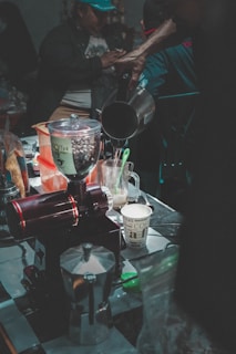 A busy coffee preparation area filled with various coffee-making tools and ingredients, including a coffee grinder, moka pot, and paper cup. A person pours liquid from a kettle into a cup. The scene is dimly lit, with a focus on the coffee process and equipment.