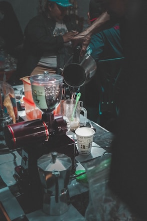 A busy coffee preparation area filled with various coffee-making tools and ingredients, including a coffee grinder, moka pot, and paper cup. A person pours liquid from a kettle into a cup. The scene is dimly lit, with a focus on the coffee process and equipment.