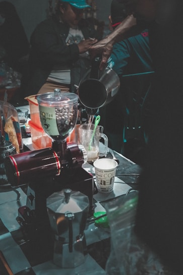 A busy coffee preparation area filled with various coffee-making tools and ingredients, including a coffee grinder, moka pot, and paper cup. A person pours liquid from a kettle into a cup. The scene is dimly lit, with a focus on the coffee process and equipment.