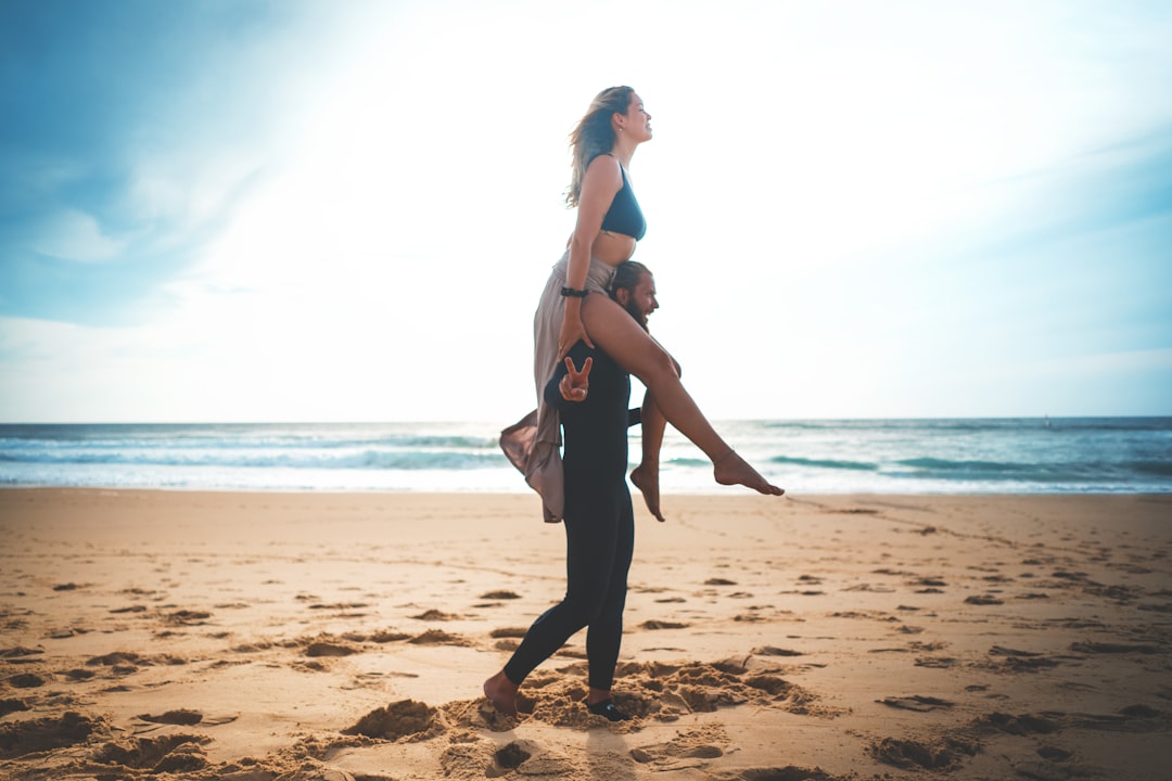 man carrying woman while walking near seashore under blue and white skies