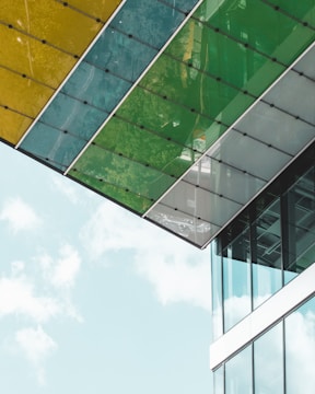 A modern architectural structure featuring large glass panels with green and yellow tinting. The building's facade reflects the surrounding environment, creating an interplay of colors and transparency. The sky is visible in the background, adding a sense of openness and light.