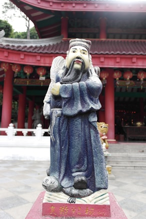 A colorful statue of a bearded figure, possibly a deity or scholar, stands with an expression of authority. The figure wears a detailed robe in shades of blue and is positioned on a platform with inscriptions. Behind the statue is a traditional Chinese architectural structure with red pillars and decorative lanterns, adding to the cultural ambiance.