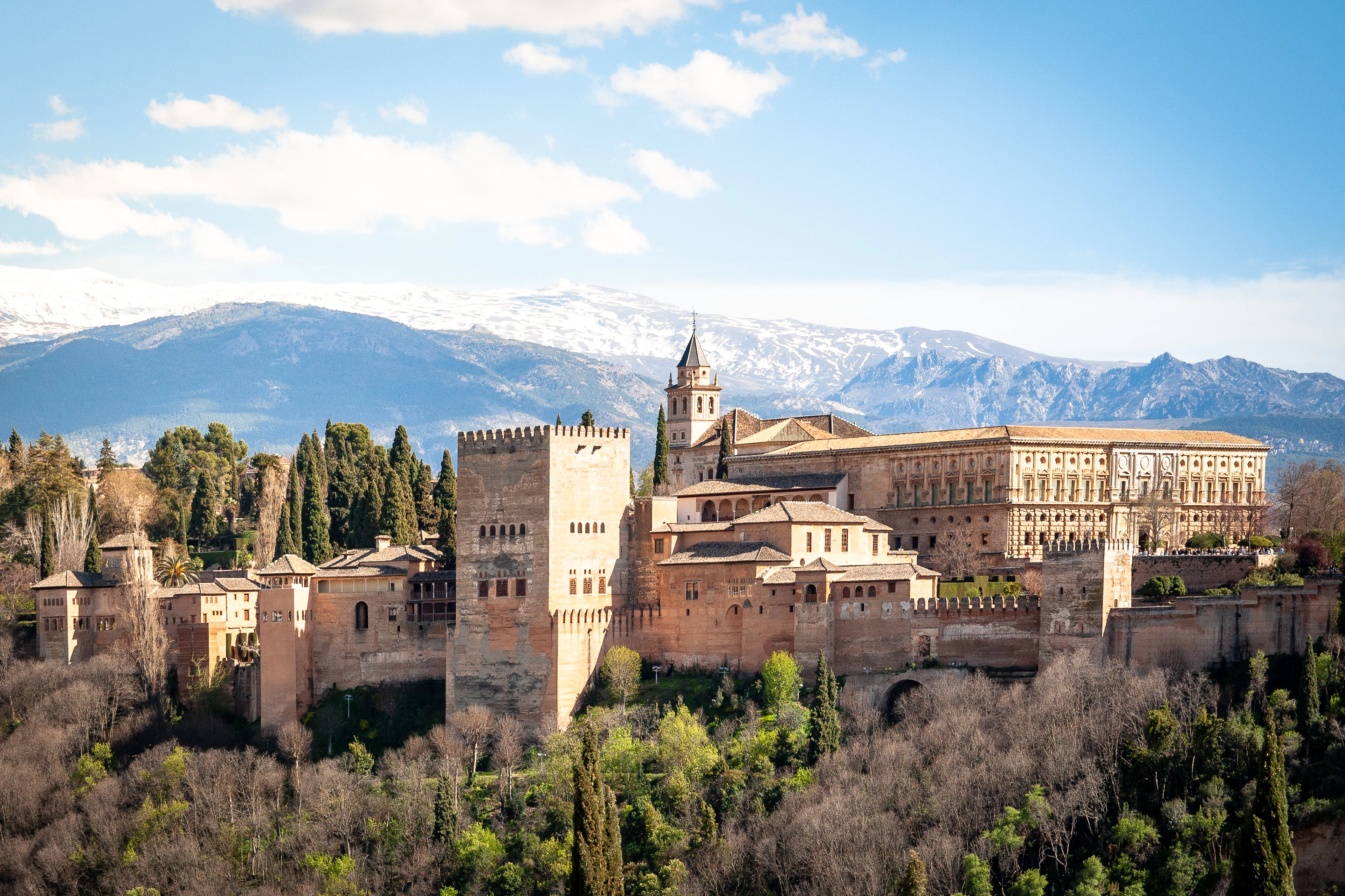 The Alhambra palace and its towers rising above wooded slopes in Granada, with the snow-capped Sierra Nevada mountains behind