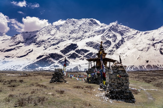 A serene landscape featuring a small stone structure adorned with colorful prayer flags in the foreground, set against a backdrop of majestic snow-covered mountains under a clear blue sky.