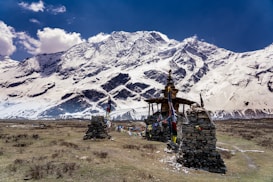 A serene landscape featuring a small stone structure adorned with colorful prayer flags in the foreground, set against a backdrop of majestic snow-covered mountains under a clear blue sky.