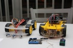 Two small wheeled robotic platforms with chassis and colorful wiring are placed on a table. Each robot features a yellow base with black tires and exposed circuit components. Nearby, a blue and black electronic board is connected with a wire.