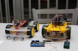 Two small wheeled robotic platforms with chassis and colorful wiring are placed on a table. Each robot features a yellow base with black tires and exposed circuit components. Nearby, a blue and black electronic board is connected with a wire.