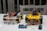 Two small wheeled robotic platforms with chassis and colorful wiring are placed on a table. Each robot features a yellow base with black tires and exposed circuit components. Nearby, a blue and black electronic board is connected with a wire.