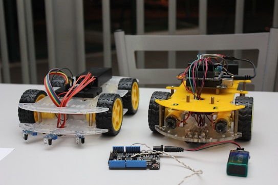 Two small wheeled robotic platforms with chassis and colorful wiring are placed on a table. Each robot features a yellow base with black tires and exposed circuit components. Nearby, a blue and black electronic board is connected with a wire.