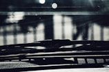 The image captures a close-up view of a car windshield and dashboard. The focus is on the windshield wipers with a blurred background showing some light reflections. The image has a dark and shadowy ambiance, with some light filtering through.